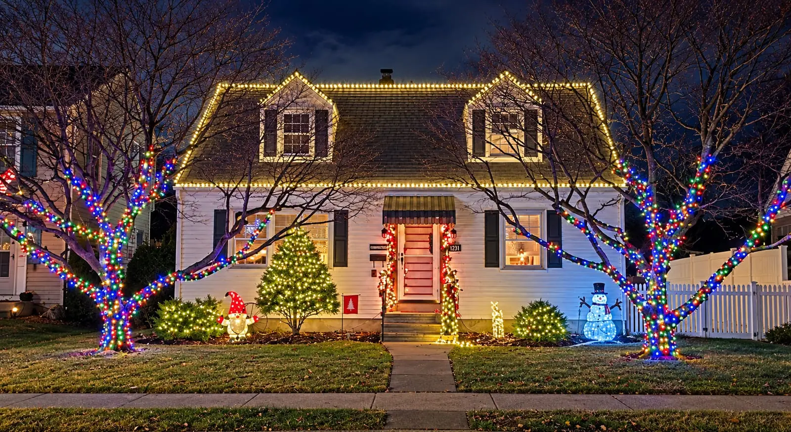 SZNS holiday lighting on a white Cape Cod home in Bethlehem, PA, with warm-white lights outlining the roofline and dormers and multicolor lights wrapped up the front-yard trees.